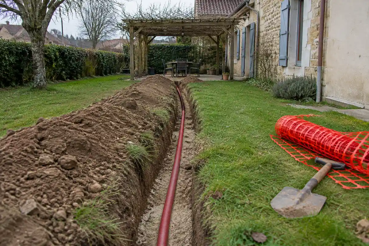 Tranchée dans une pelouse vers une pergola, gaine électrique rouge au fond et rouleau de grillage avertisseur rouge à côté